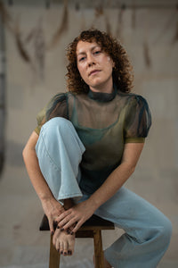 Woman sitting on a stool wearing a sheer top and light blue jeans against a neutral background
