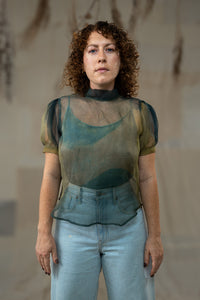 Woman wearing a sheer green top and light blue jeans against a neutral background