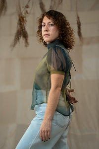 Woman wearing a sheer green top and light blue jeans against a neutral background
