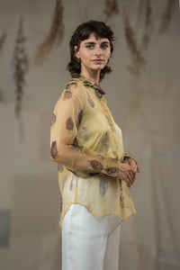 A female model wearing HeenaAgrima's tannin silk organza shirt, a beige organza sheer shirt with a floral pattern, featuring a round neckline and long sleeves, paired with white trousers.