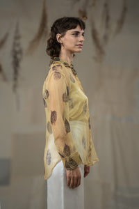 A female model wearing HeenaAgrima's tannin silk organza shirt, a beige organza sheer shirt with a floral pattern, featuring a round neckline and long sleeves, paired with white trousers.