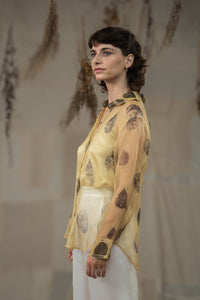 A female model wearing HeenaAgrima's tannin silk organza shirt, a beige organza sheer shirt with a floral pattern, featuring a round neckline and long sleeves, paired with white trousers.