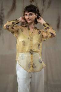 A female model wearing HeenaAgrima's tannin silk organza shirt, a beige organza sheer shirt with a floral pattern, featuring a round neckline and long sleeves, paired with white trousers.