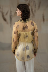 A female model wearing HeenaAgrima's tannin silk organza shirt, a beige organza sheer shirt with a floral pattern, featuring a round neckline and long sleeves, paired with white trousers.