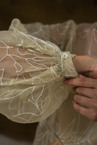 Cloase-up of a hand of a Woman wearing HeenaAgrima's Yellow Eve Silk Organza Top, a sheer top with leaf patterns and yellow pants against a neutral background. 