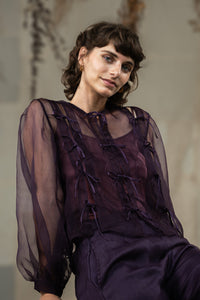 Woman wearing HeenaAgrima's Aubergine Zoe Silk Organza Top, a sheer purple blouse with ruffled details against a neutral background. 
