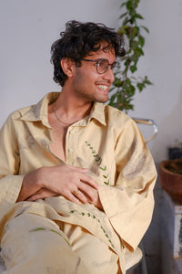 Man wearing a light-colored embroidered shirt sitting indoors with plants in the background