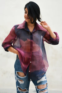 Woman wearing a sheer ombre dyed silk shirt over a bustier top with ripped jeans against a white background