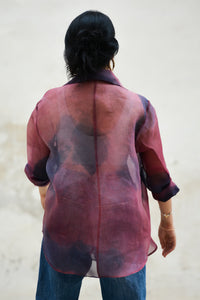 Person wearing a sheer pink and purple jacket over a dark top and jeans on a light background