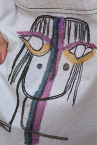 Close-up of a Woman wearing HeenaAgrima's Nina Cotton Shirt, a white shirt with colorful face designs, holding sunglasses.