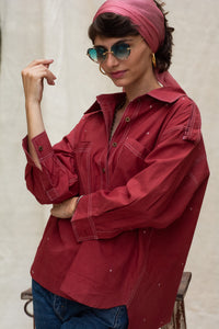 Woman wearing a red organic cotton shirt, named cherry, made by HeenaAgrima, blue glasses and pink headscarf against a plain background. 