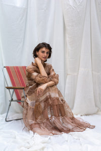 Woman sitting and Wearing HeenaAgrima's Mocha Dress, a Sheer, tiered dress with a brown and beige color scheme on a white background. 