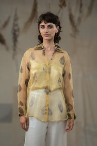 A female model wearing HeenaAgrima's tannin silk organza shirt, a beige organza sheer shirt with a floral pattern, featuring a round neckline and long sleeves, paired with white trousers.
