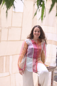 Woman wearing HeenaAgrima's rainbow rays organza shirt, a colorful shirt sitting outdoors with a light-colored wall and greenery in the background.