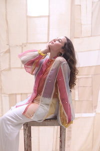 Woman wearing HeenaAgrima's rainbow rays organza shirt, a colorful shirt with little hearts, sitting outdoors with a light-colored wall and greenery in the background.