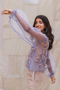 Woman wearing HeenaAgrima's Zoe Lilac Top a sheer top with lace details in front and back of light curtains. 