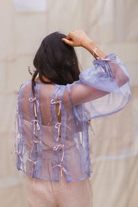 Woman wearing HeenaAgrima's Zoe Lilac Top a sheer top with lace details in back of light curtains