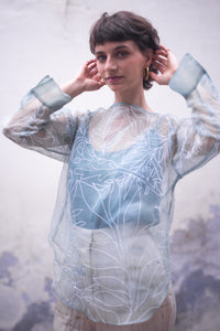 Woman wearing a sheer blue top with floral patterns against a blurred background - HeenaAgrima