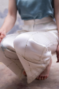 woman sitting while wearing HeenaAgrima's light-colored checkered satin silk pants against a quilt with a blurred background
