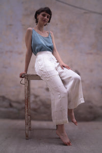 Woman sitting on a chair wearing a blue top and HeenaAgrima's light-colored checkered satin silk pants against a textured wall.