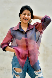 Woman wearing a sheer pink and purple blouse over a bustier  top with ripped jeans, smiling against a plain background.